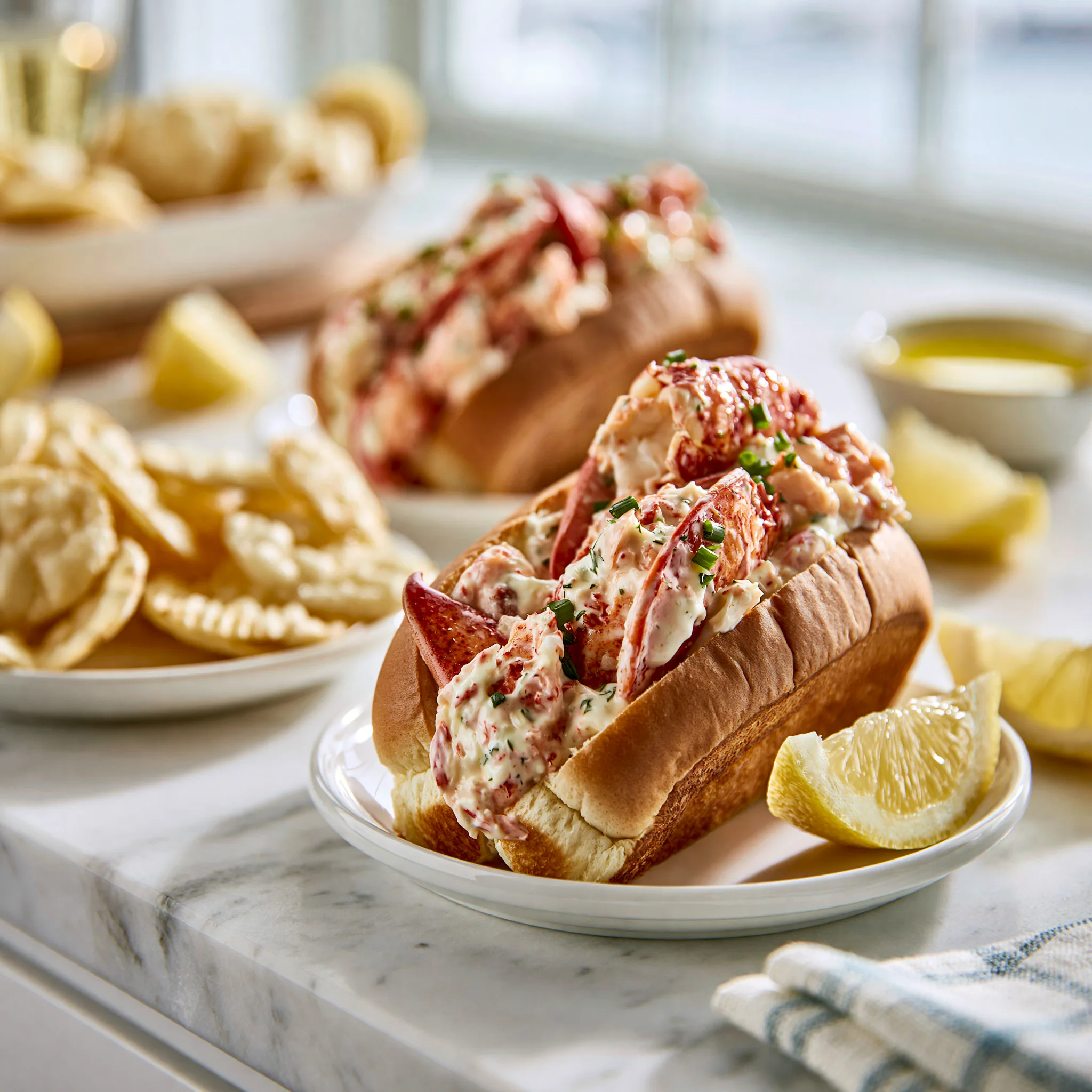 New England lobster roll served on a white plate with a slice of lemon with dip blurred in the background.